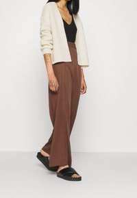 Knit cream cardigan over a black top, paired with loose brown trousers and black slide sandals. Features include relaxed design and textured fabric.