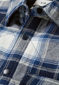 Rutigt skjorta i blå, vit och grå färg. Tillverkad av ett mjukt tyg, med knäppning fram och vikta kragar. Mörka knappar synliga.