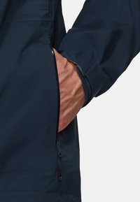Veste imperméable marine avec une poche latérale. Présente une texture lisse et une fermeture éclair sur la poche. Les poignets sont ajustables.
