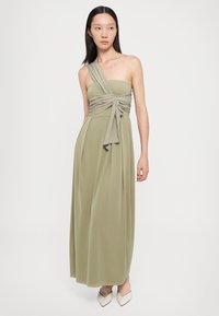 Rochie maxi verde măsliniu cu un design cu un umăr, detaliu nod în zona bustului, siluetă fluidă și textură netedă. Asortată cu pantofi cu toc crem.