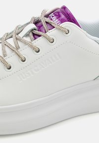 Witte sneaker met reliëf "JUSTCAVALLI" logo, zilveren veters en paarse glitters op de tong. Glad synthetisch materiaal, rubberen zool.
