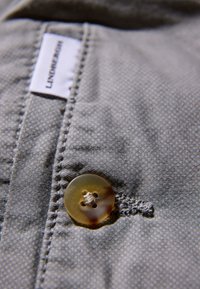 Grauer Stoff mit strukturierter Webart, versehen mit einem großen, durchscheinenden Knopf mit vier Löchern, der mit sichtbarem Faden angenäht ist.