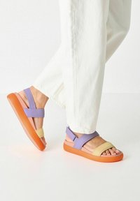 Multicolored sandals with purple, yellow, and orange straps, a wide cushioned sole, and a smooth texture; designed for comfort and style.