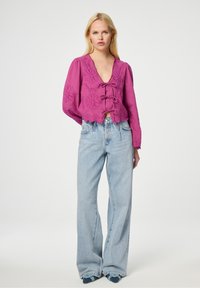 Roze cropped blouse met bloem borduurwerk, diepe V-hals en sluiting met een strik aan de voorkant. Gecombineerd met lichtblauwe, wijde jeans en blauwe instappers.