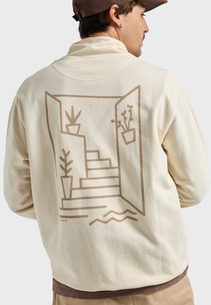 Man wearing beige long-sleeve shirt with brown line art of stairs and potted plants on its back, brown cap, and beige pants.