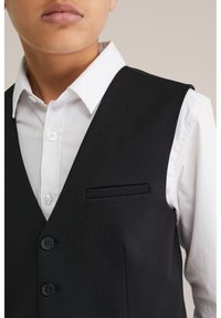 Černá vesta s texturovanou tkaninou, s jednou kapsou na hrudi, dvěma knoflíky a pod ní bílá košile s límečkem.