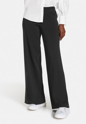 Osoba ubrana w czarne spodnie o szerokich nogawkach, białe trampki i białą koszulę z długim rękawem i bufkami.