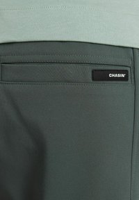Pantaloni in tessuto verde con vestibilità slim, un taschino sul retro e un'etichetta rettangolare nera con la scritta "CHASIN'". Texture liscia e design sartoriale.