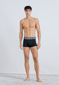 Czarne bawełniane bokserki z białą tasiemką "CALVIN KLEIN", prezentujące dopasowany krój na męskim modelu na jednolitym tle.