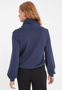 Nachtblauer Pullover mit hohem Kragen und Ballonärmeln, aus weichem Stoff gefertigt. Die Rückseite weist ein glattes Design und eine lockere Passform auf.