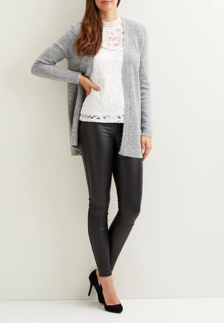 Femme portant des talons hauts noirs, des leggings en cuir noirs, un haut en dentelle blanche et un cardigan gris tricoté, debout contre un mur blanc.