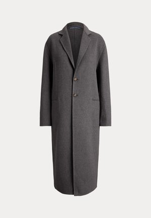 Femme portant un manteau gris foncé, un col roulé, un pantalon large, des lunettes de soleil noires, tenant un sac à main en cuir noir, debout devant un fond neutre.