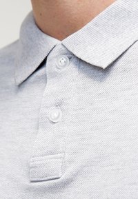 Harmaa polo-paita, jossa on teksturoitu kangas, kauluspääntie ja kolme valkoista nappia etuosassa.