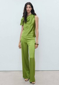Haut en satin vert avec une volute sur une épaule, associé à un pantalon palazzo assorti. Texture lisse, design épuré et sandales neutres.