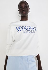 Bela pulover s povezanimi modrimi vezenimi besedami "MYKONOS BLUE OCEAN." Mehka tkanina, sproščen kroj, s spuštenimi rameni. Pod njim svetlomodre hlače.