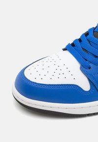 Blå lädersneakers med vit tåskydd, perforeringar, svarta snören och texturerade blå detaljer. Gummisula synlig i botten.