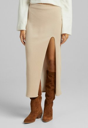 Jupe midi en maille côtelée beige avec une fente devant, portée avec des bottes hautes en daim marron et un haut blanc à manches longues.