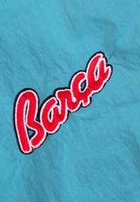 Turkoois stof met een opvallende rode geborduurde tekst "Barca," omlijnd in zwart. Het oppervlak heeft een gestructureerd uiterlijk.