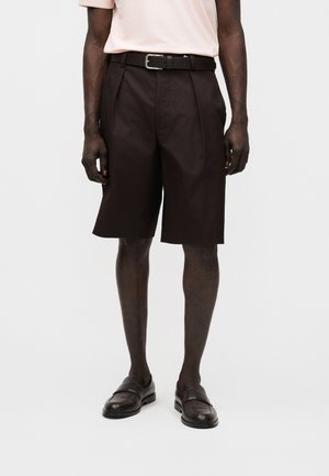 Mees, kes kannab musti põlveni pükse musta vööga, kahvatu roosa särgi ja mustade nahast slip-on kingadega, seisab ühevärvilise tausta ees.