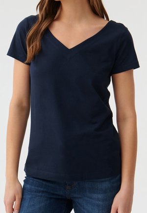Femme portant un T-shirt bleu marine à manches courtes et col en V, associé à un jean bleu sur fond blanc uni.