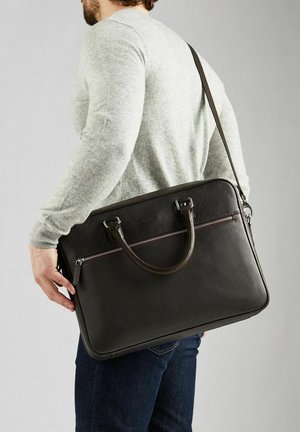 Mies kantaa mustaa nahkaista salkkua, jossa on olkahihna ja kädensijat, pukeutuneena harmaaseen neulepaitaan ja tummansinisiin farkkuihin, selin katsojaan.