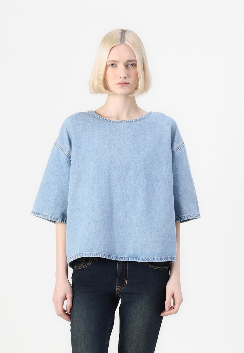 Top in denim blu chiaro, corto, con maniche corte, scollatura rotonda e dettagli a orlo grezzo. Abbinato a jeans in denim scuro. Texture liscia.