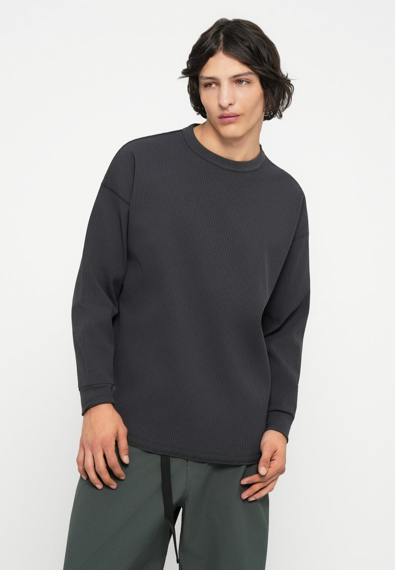 T-shirt à manches longues gris foncé côtelé avec un col rond, présentant une finition texturée et une coupe décontractée. Assorti avec un pantalon vert.