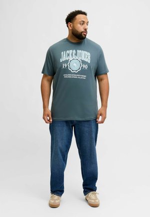 Dark teal cotton t-shirt with white screen-printed graphics and text. Paired with loose blue denim jeans and light brown sneakers.