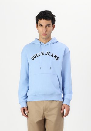 Lichtblauwe hoodie met zwarte "GUESS JEANS" tekst, trekkoorden, voorzak en ribgebreide manchetten en onderrand. Gemaakt met beige broek.