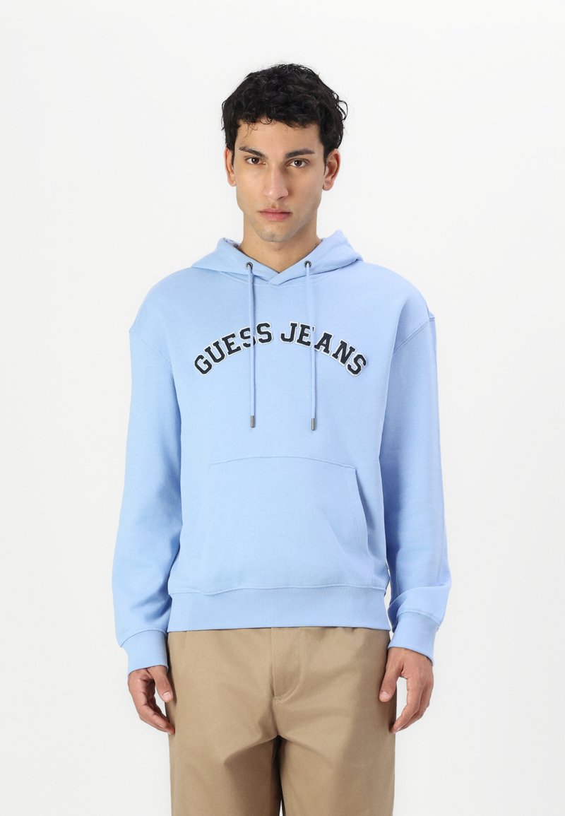 Sudadera azul clara con el texto negro "GUESS JEANS", cordones, bolsillo frontal y puños y dobladillo acanalados. Combinada con pantalones beige.