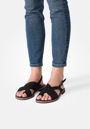 Legs wearing blue denim jeans and black flat sandals with crossed straps and ankle buckles on a white background.