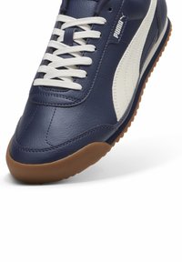 Námořnické kožené tenisky s bílými akcenty, texturovaným povrchem a světle hnědou gumovou podrážkou. Obsahují tradiční šněrování a branding Puma.