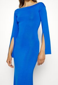 Långärmad, off-the-shoulder klänning i ljusblått, med sidofickor och en figurnära design gjord av ett smidigt, stretchigt tyg.