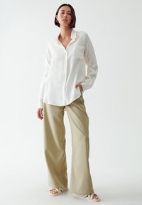Camicia bianca a maniche lunghe con colletto e taschino frontale, abbinata a pantaloni beige a vita alta e gamba larga. Tessuto morbido, vestibilità comoda.