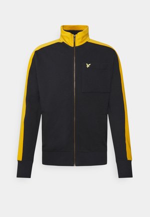 Chaqueta negra con cremallera y rayas amarillas en las mangas y el cuello, con un pequeño logo de un pájaro amarillo en el bolsillo del pecho izquierdo.