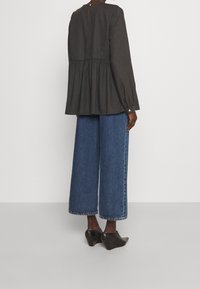 Mørkegrå peplum-bluse med lange ærmer, lukning bagpå med knapper, parret med højtaljede vide blå denim-jeans og sorte kilehæle.