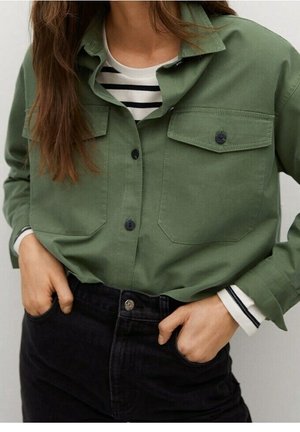 Femme portant une chemise verte boutonnée sur un haut rayé noir et blanc, les mains dans les poches d'un pantalon noir taille haute.