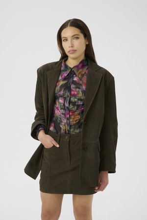 Woman with long brown hair wearing dark olive oversized blazer and skirt, colorful ruched button-up shirt, posing with one hand in pocket.