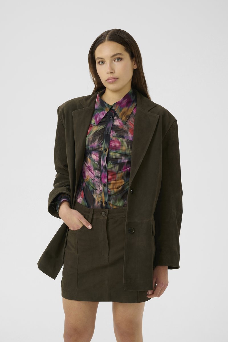 Frau mit langen braunen Haaren, die einen dunkeloliven, oversized Blazer und einen Rock trägt, sowie ein farbenfrohes, gerafftes Hemd mit Knöpfen, posiert mit einer Hand in der Tasche.