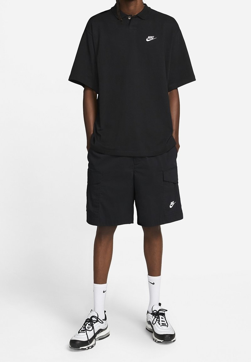 Polo noir à manches courtes avec un logo Nike, associé à un short cargo noir avec des poches latérales et un détail de logo, complété par des baskets noir et blanc.