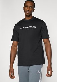 Μαύρο βαμβακερό μπλουζάκι με λευκό λογότυπο "JUVENTUS". Μανίκια κοντά, λαιμόκοψη στρογγυλή, με άνετη εφαρμογή. Φορεμένο με γκρι αθλητική φόρμα.