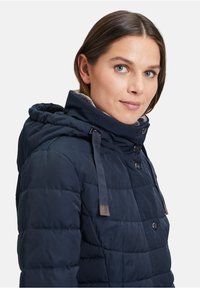 Marineblauwe gewatteerde jas met een trekkoord capuchon, knopen aan de voorkant en grijze fleece voering. Zachte textuur en gestikt patroon.