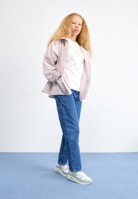 Světlounce růžová bunda na zip přes bílé tričko s grafickými potisky, doplněná modrými džínami a šedými teniskami. Hladké textury a pohodlný střih.