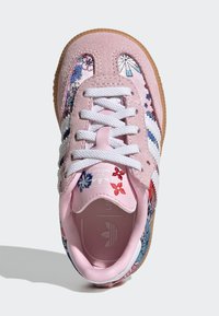 Zapatilla deportiva de color rosa claro con patrón floral en los lados, cordones blancos, tres rayas blancas y una suela de goma color beige. Materiales lisos y texturizados.