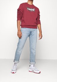 Sudadera de forro polar burdeos con el logotipo negro de "Levi's", jeans rectos de color azul claro y zapatillas blancas con acentos rojos y detalles en negro.