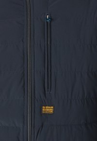 Tamsiai mėlyna pamušta striukė su horizontaliomis siūlėmis, užtrauktuku užsegama krūtinės kišenė ir G-Star Originals logotipo etikete. Lygus paviršius, standartinė forma.
