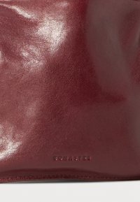 Burgundská kožená kabelka s lesklým povrchom, ktorý má tonálne prešívanie a embosovanú značku na spodnej strane. Hladký povrch a zjednodušený tvar.