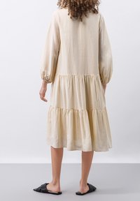 Un vestido beige a la altura de la rodilla con mangas largas estilo globo, con niveles fruncidos y un ajuste holgado. Textura suave y diseño minimalista, combinado con sandalias negras.