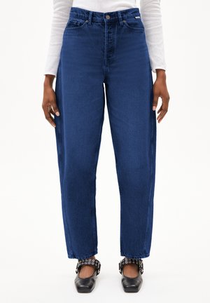 Jeans droits taille haute bleus avec un minimum de distorsion, finition en denim texturé et deux poches pour les mains. Portés avec des chaussures noires présentant des éléments en argent.