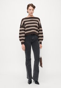 Woman wearing a brown and beige striped sweater, black high-waisted jeans, black pointed boots, and holding a small brown fringed bag.